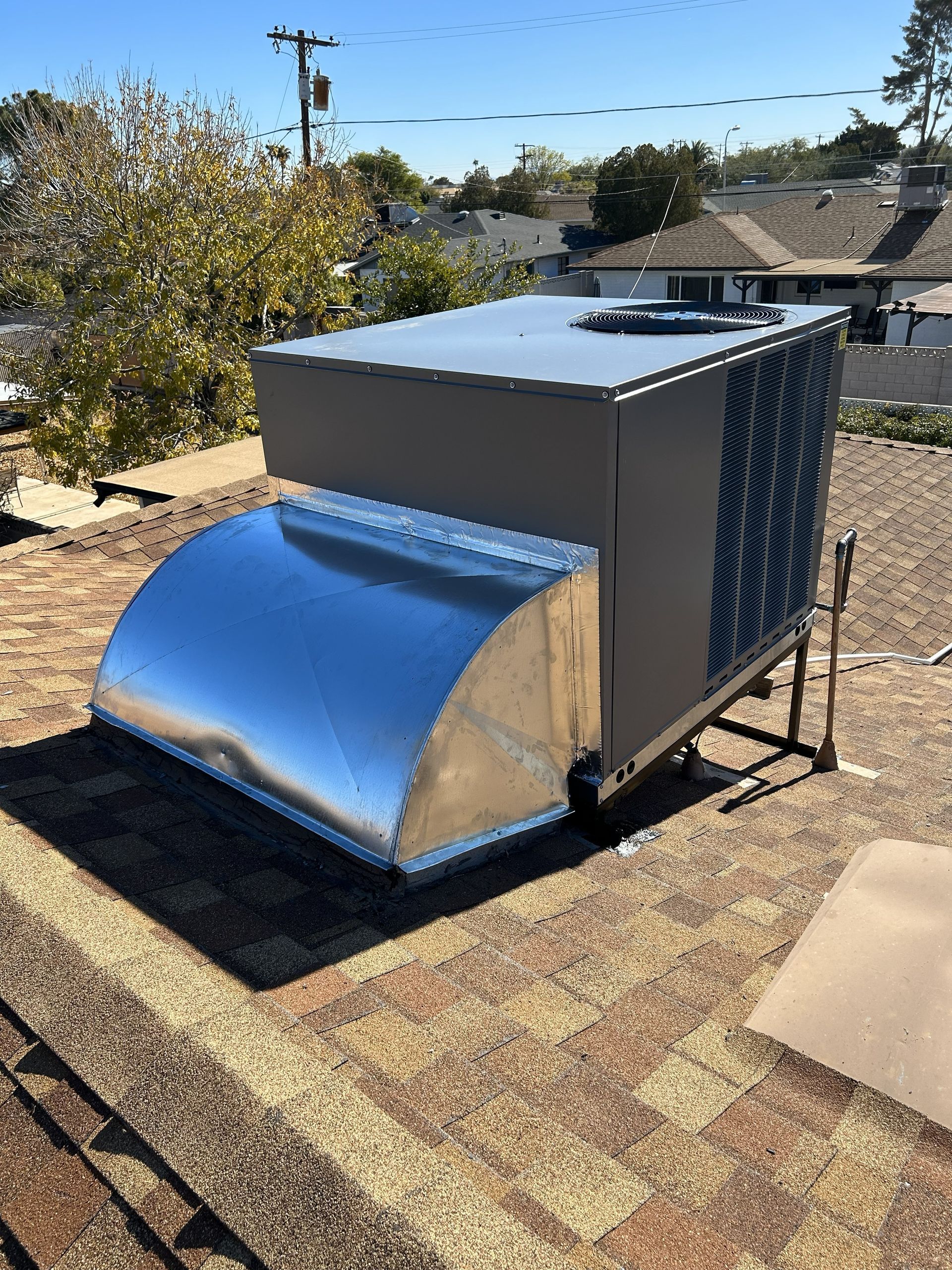 HVAC unit with silver vent hood on a rooftop with brown shingles under a blue sky.