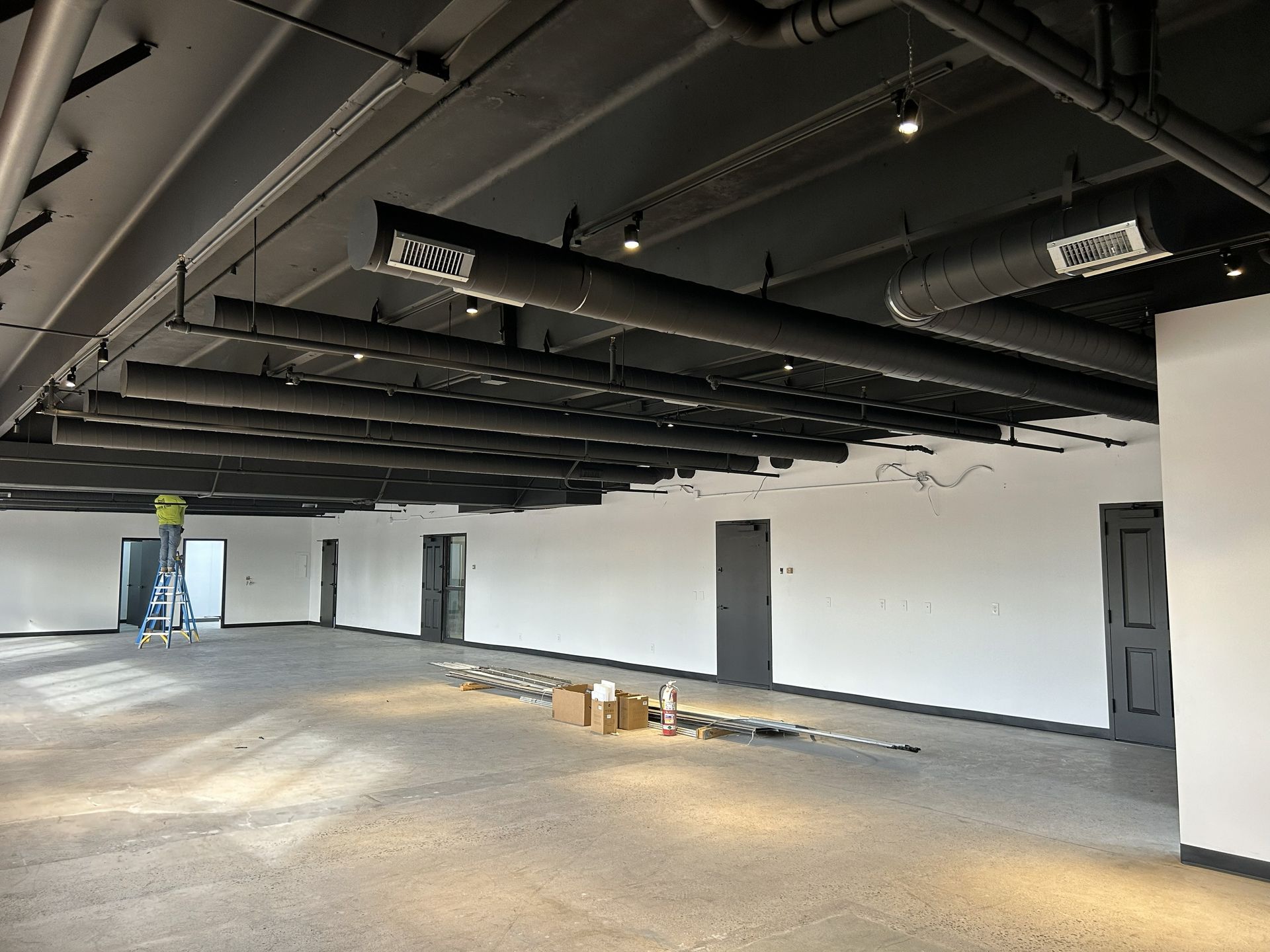 Empty room under construction, black ceiling with vents and pipes, white walls, concrete floor.