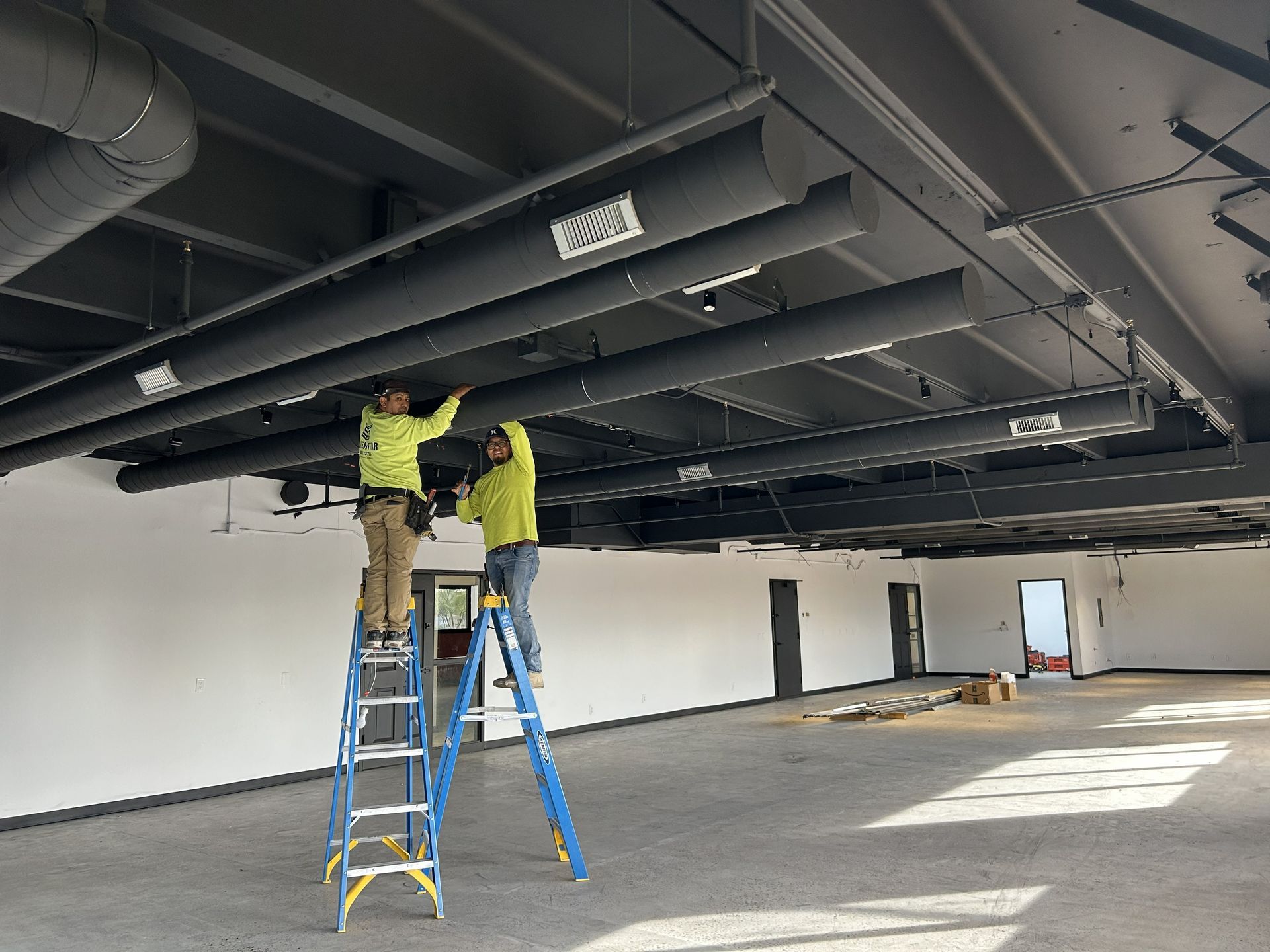Two workers on ladders installing dark-colored ceiling fixtures in an open, unfinished room.
