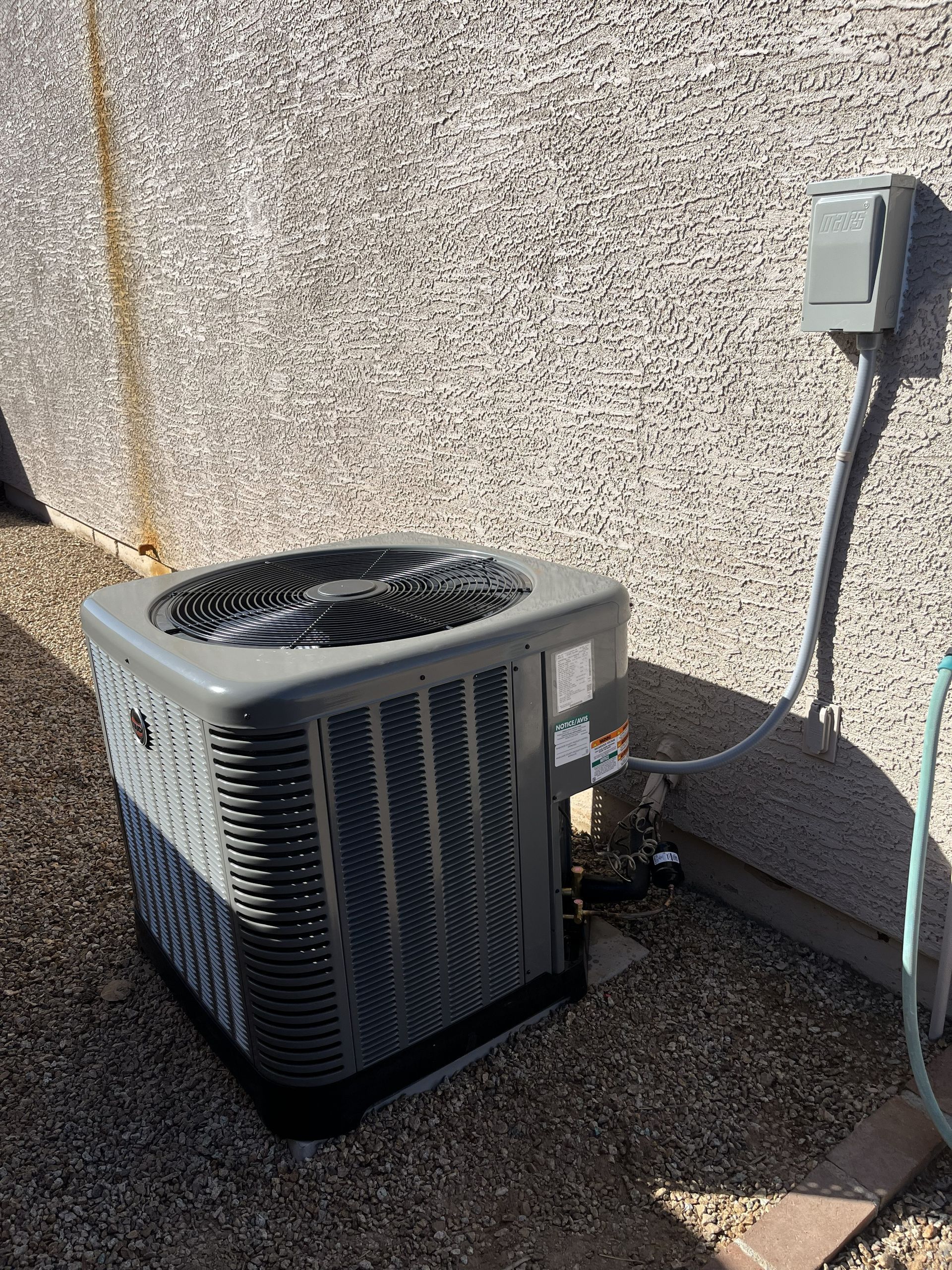 Air conditioning unit against a stucco wall, connected to power supply, sitting on gravel.