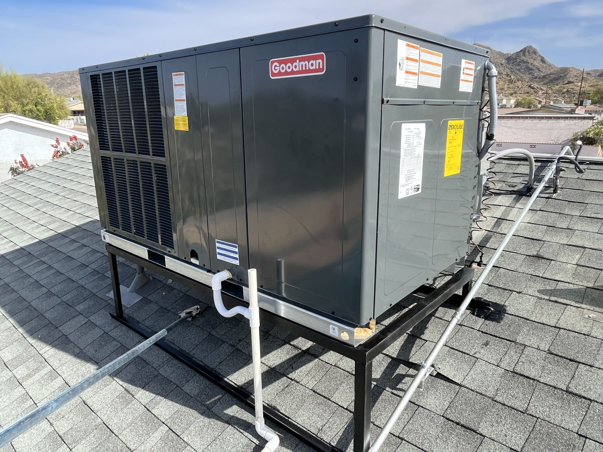 A Goodman air conditioning unit sits on a black metal platform on a rooftop.