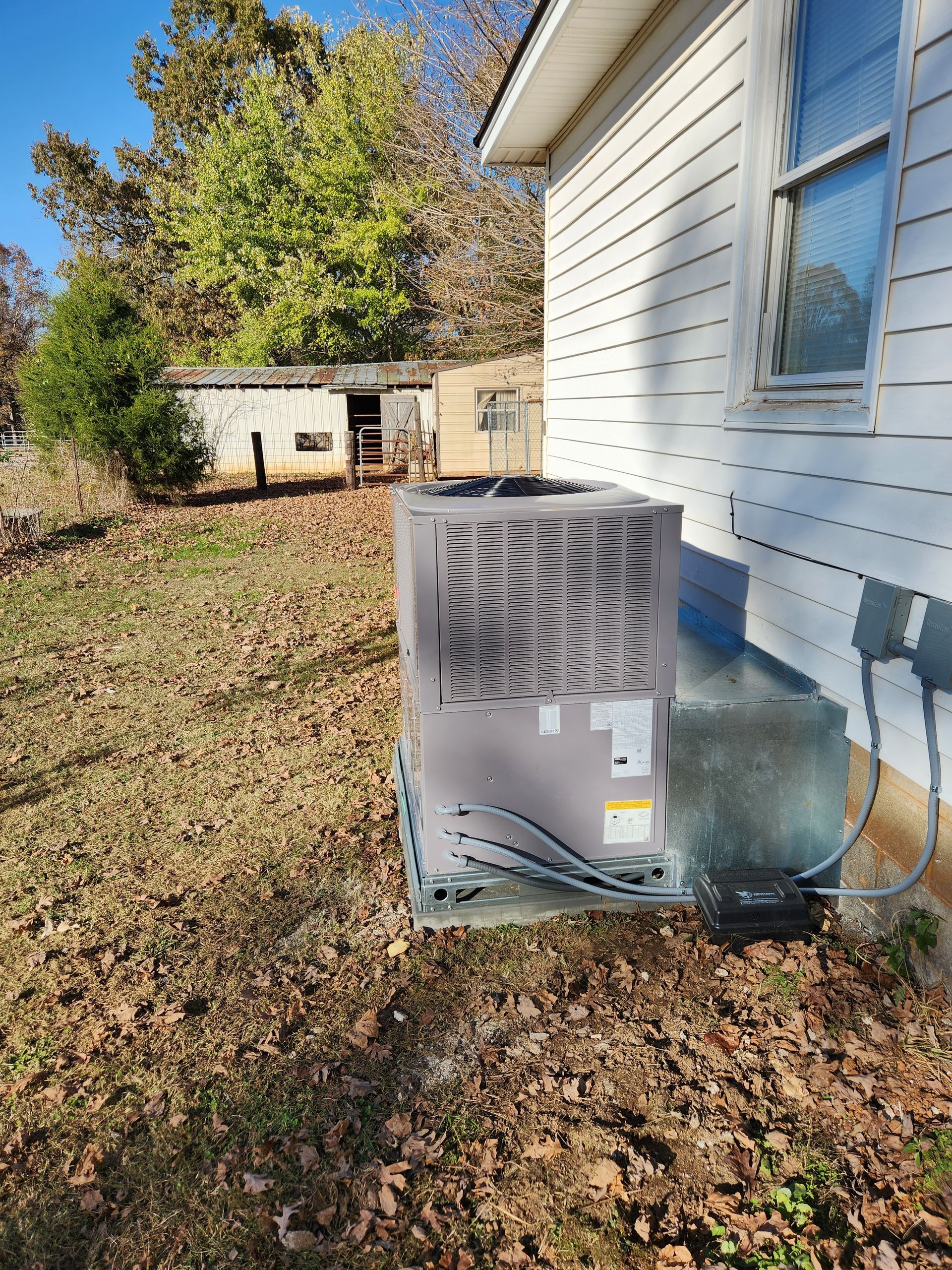 An air conditioning unit sits outside a white house, next to a shed, on a grassy yard.