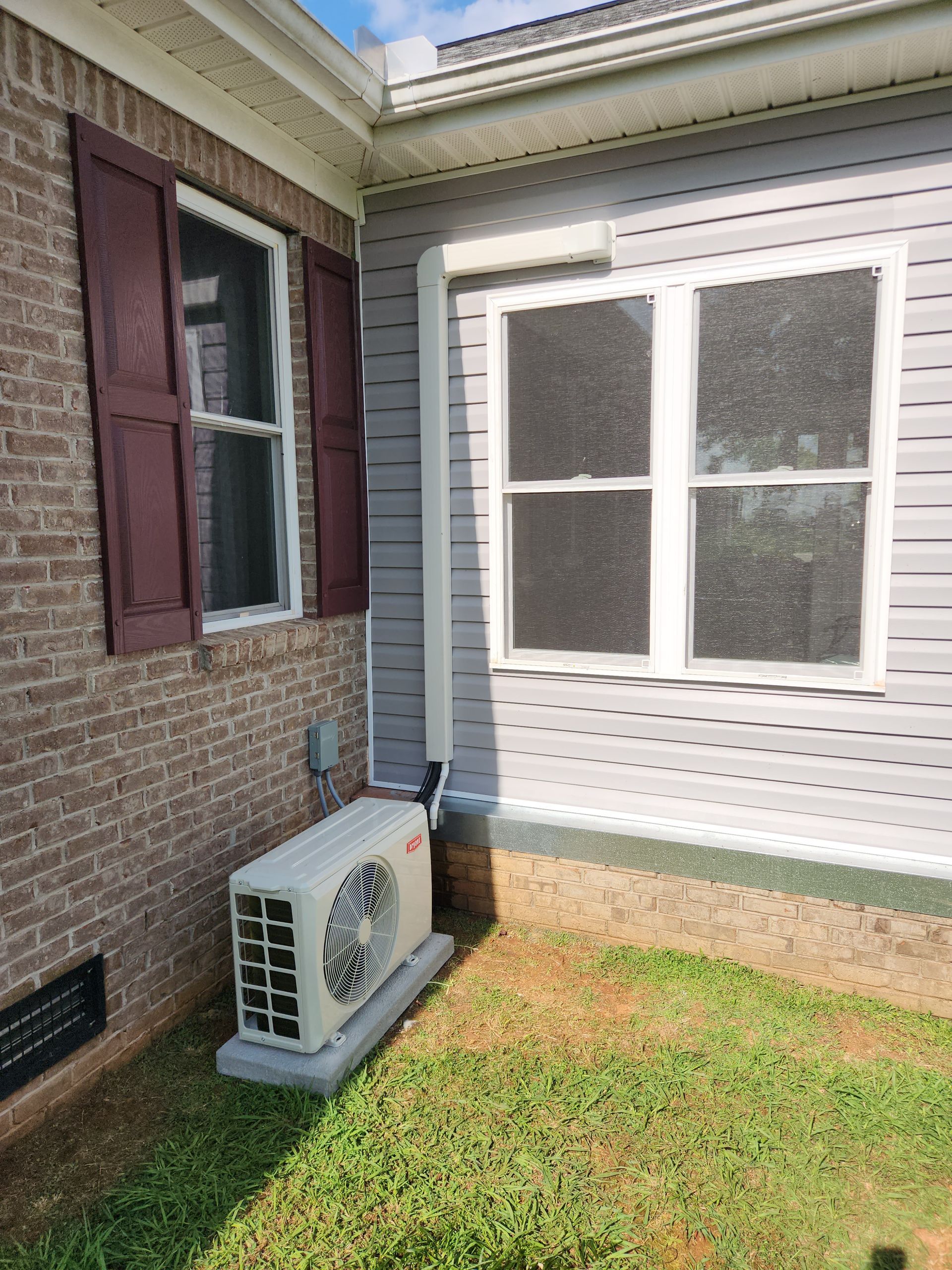 Air conditioning unit outside brick and gray siding building; brown shutters, grass.