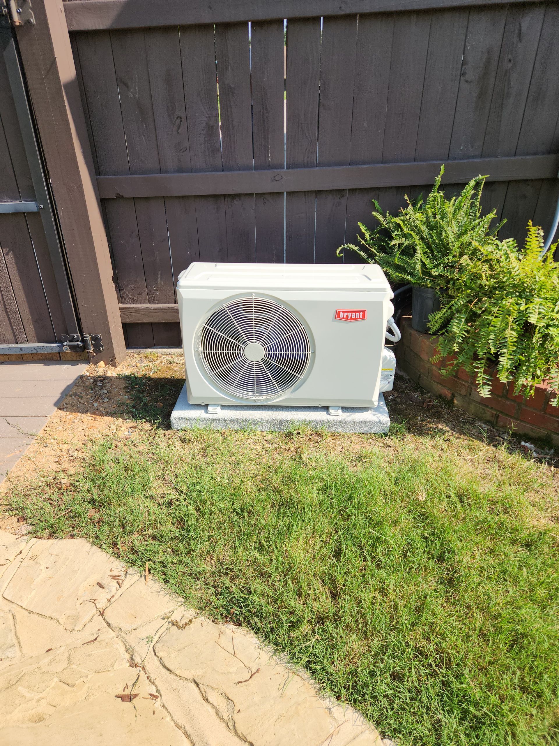White Bryant air conditioner unit on a concrete pad in a yard, near a wooden fence and green plants.