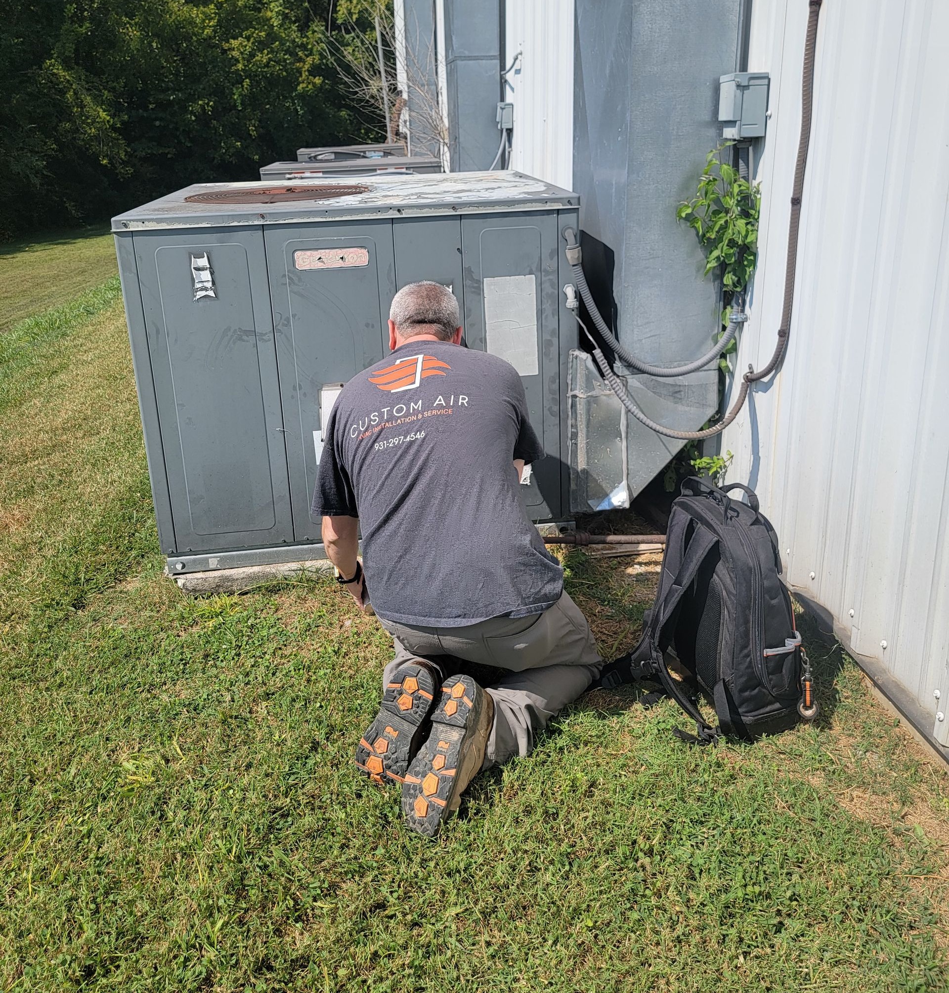 A person in work clothes kneels, inspecting an outdoor HVAC unit next to a building.