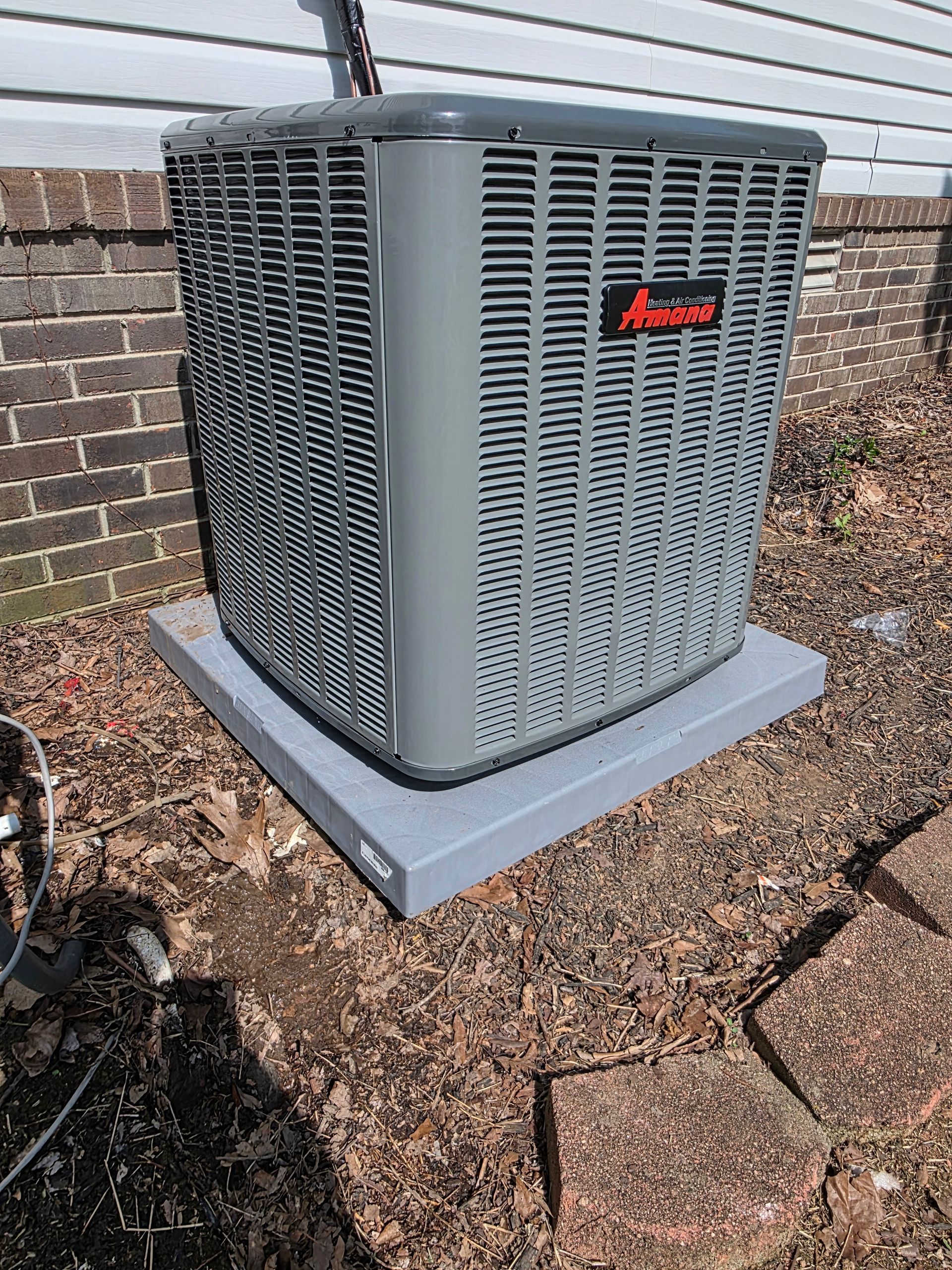 An Amana air conditioning unit sits on a concrete pad next to a brick wall.