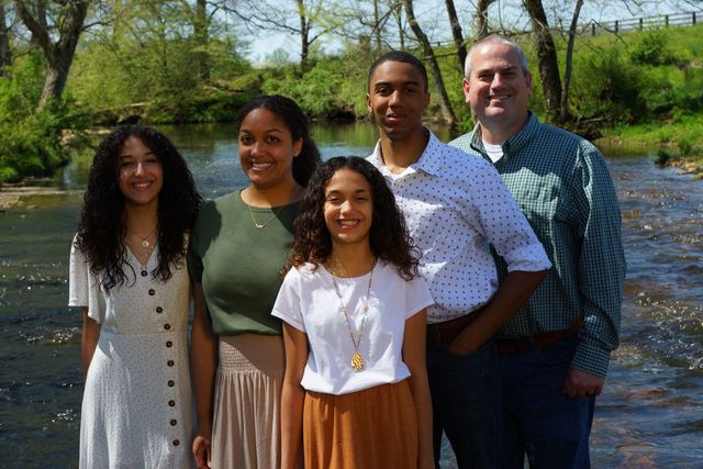 Family of five smiles by a stream; two girls in front, a man and woman, and a man stand behind.