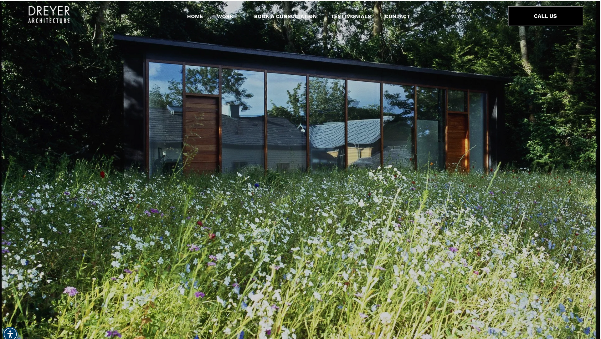 Modern glass-walled building reflects trees and a house, set in a field of wildflowers.