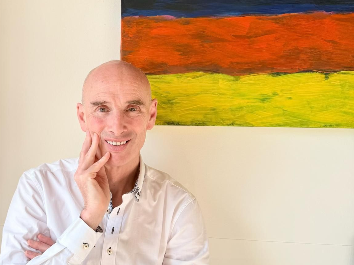 Bald man with hand on shoulder smiles in front of a colorful, dotted abstract painting.
