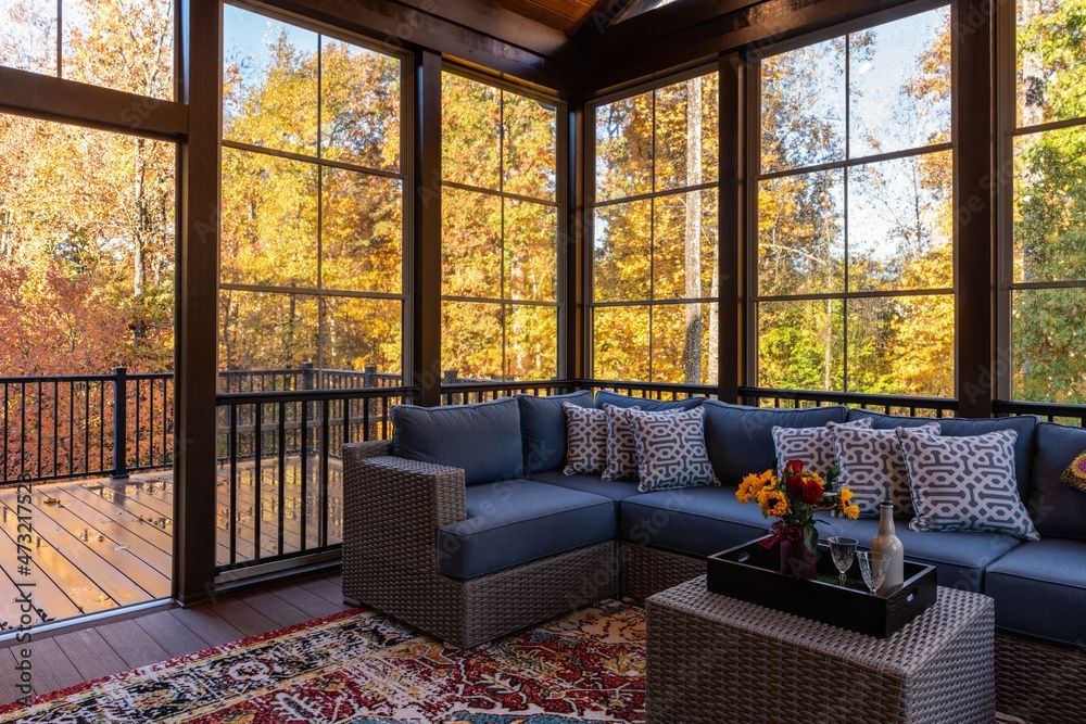 Véranda fermée avec canapé d'angle bleu, tapis et table basse ; vue sur des arbres aux feuilles dorées.