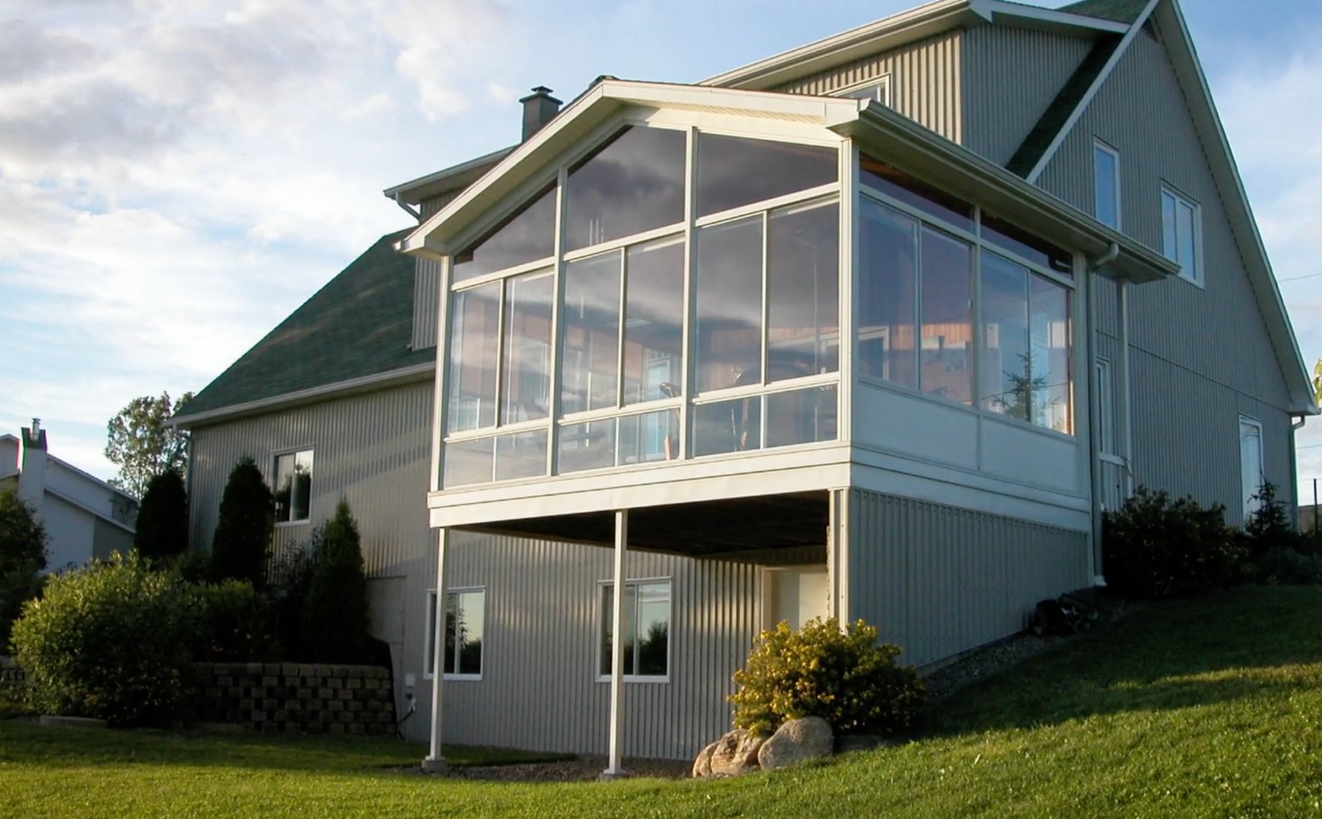 Une véranda de couleur claire avec des murs en verre attachée à une maison à deux étages, sur une colline herbeuse.