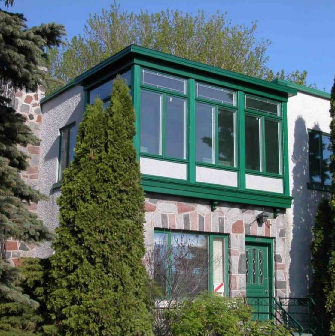 Maison en pierre avec bordure verte autour des fenêtres et de la porte ; grands arbres verts devant.