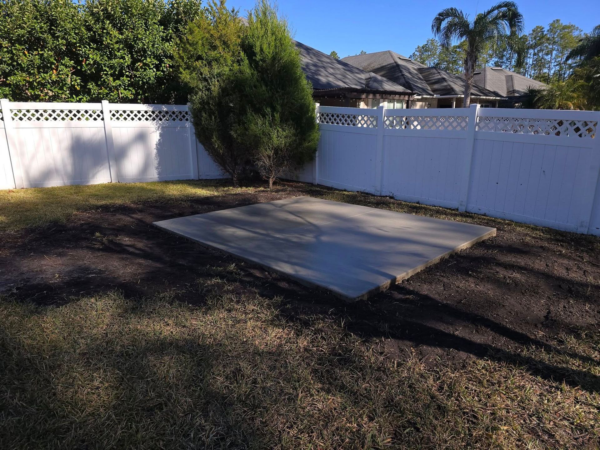 Backyard with white fence, concrete slab, and small evergreen tree.