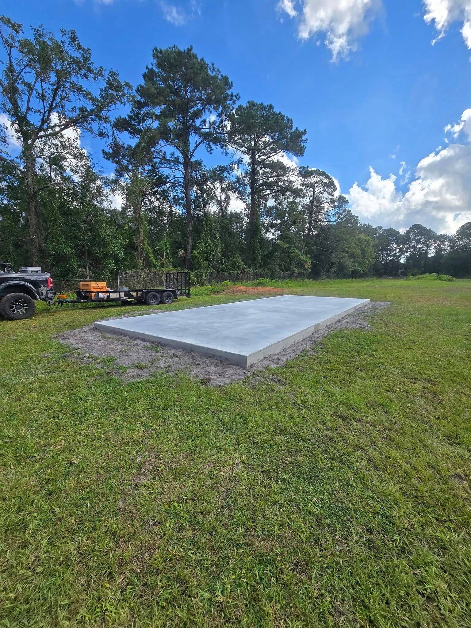 Concrete slab on a grassy field, trees in the background, blue sky, and a small trailer.