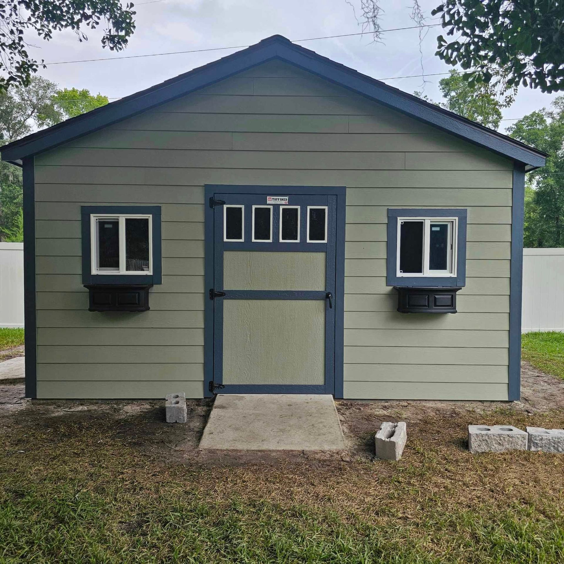 Green shed with blue trim, door, and windows. Two windows with black flower boxes. Gray concrete path.