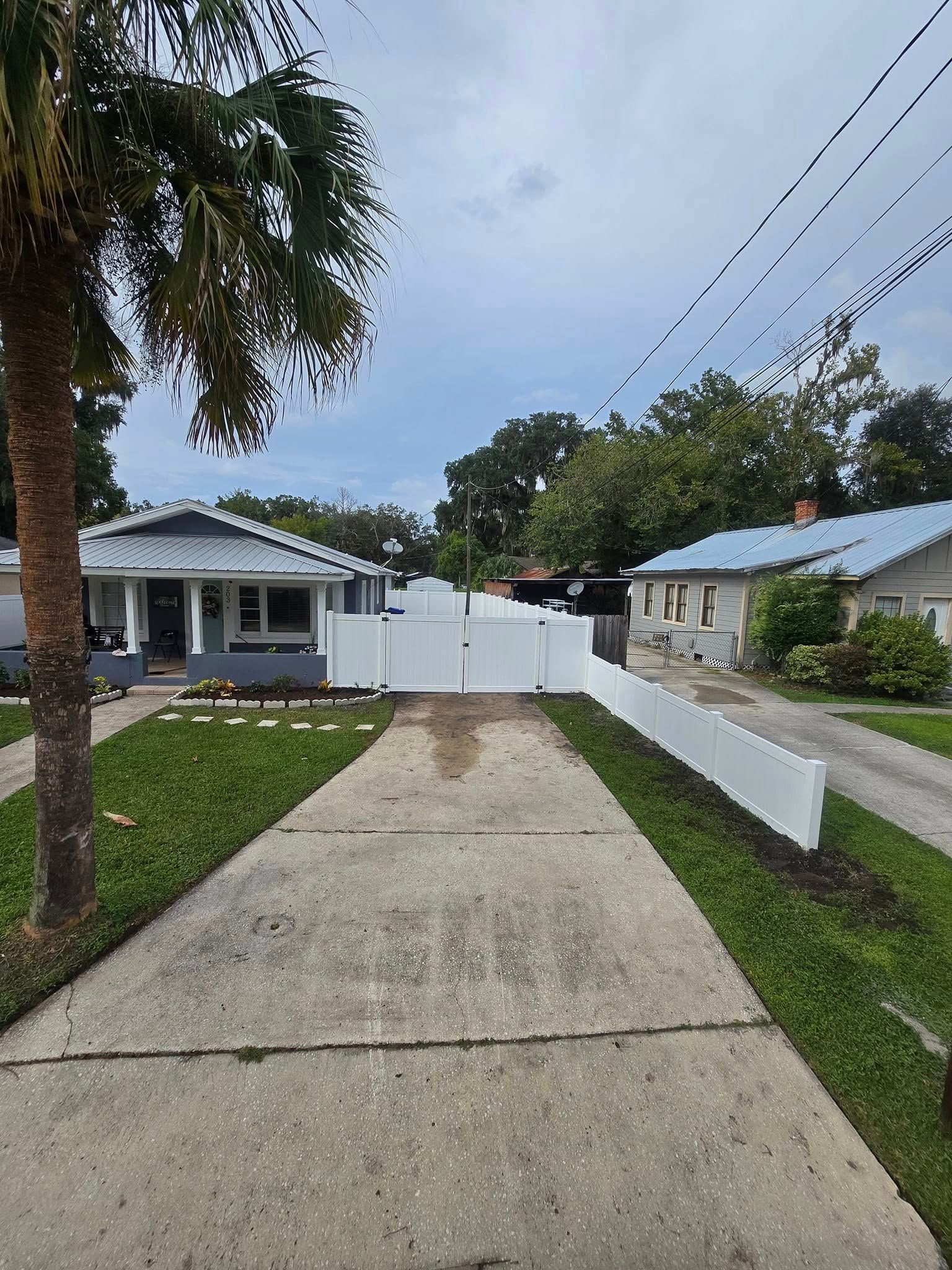 Driveway between two houses with white fences. Green grass and palm tree in view.