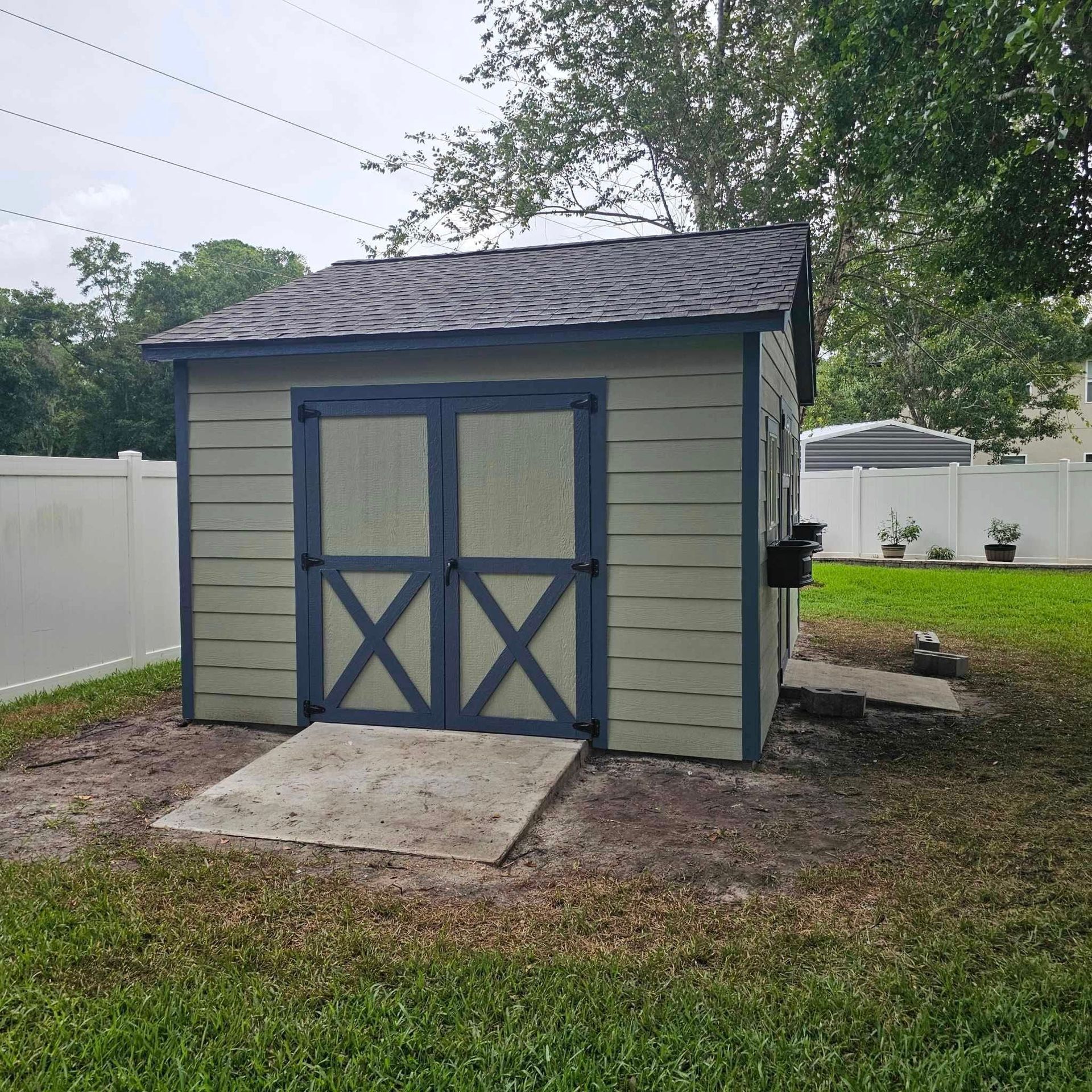 A small, green shed with blue trim and doors in a grassy yard. Concrete pad in front.