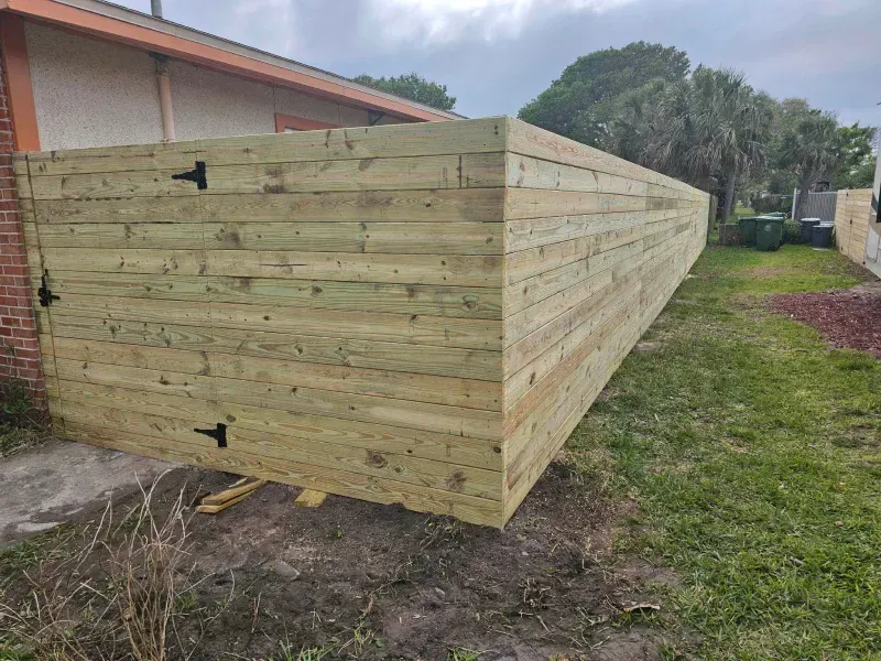 A long, unfinished wooden fence surrounding a grassy yard next to a brick building.