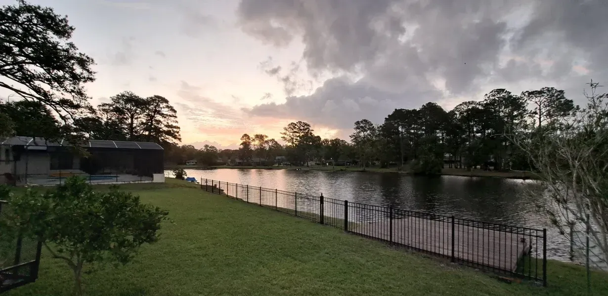 A lake at dusk with dark water, a grassy lawn, trees, and cloudy sky. A fence runs along the water.