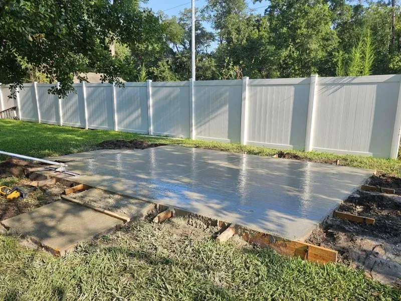 Newly poured concrete patio in a backyard with a white fence.