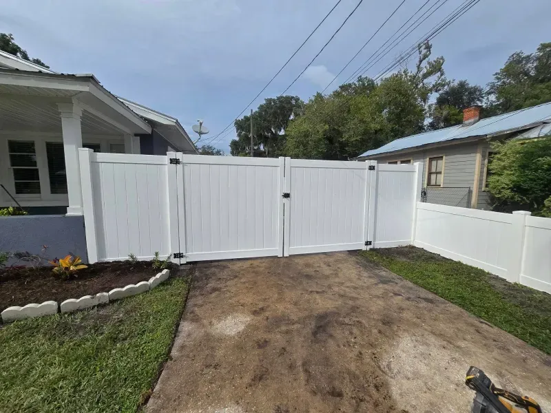 White vinyl fence with double gates in front of a driveway, in front of a house.