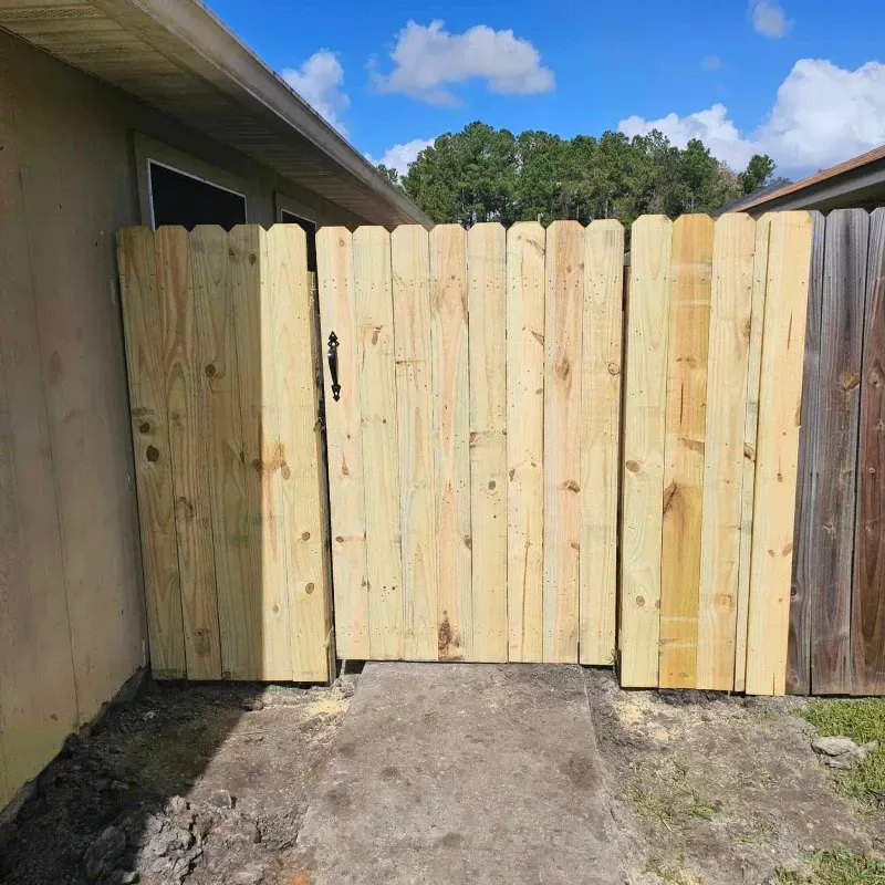 Wooden fence and gate, tan and weathered. Entrance to yard with blue sky background.