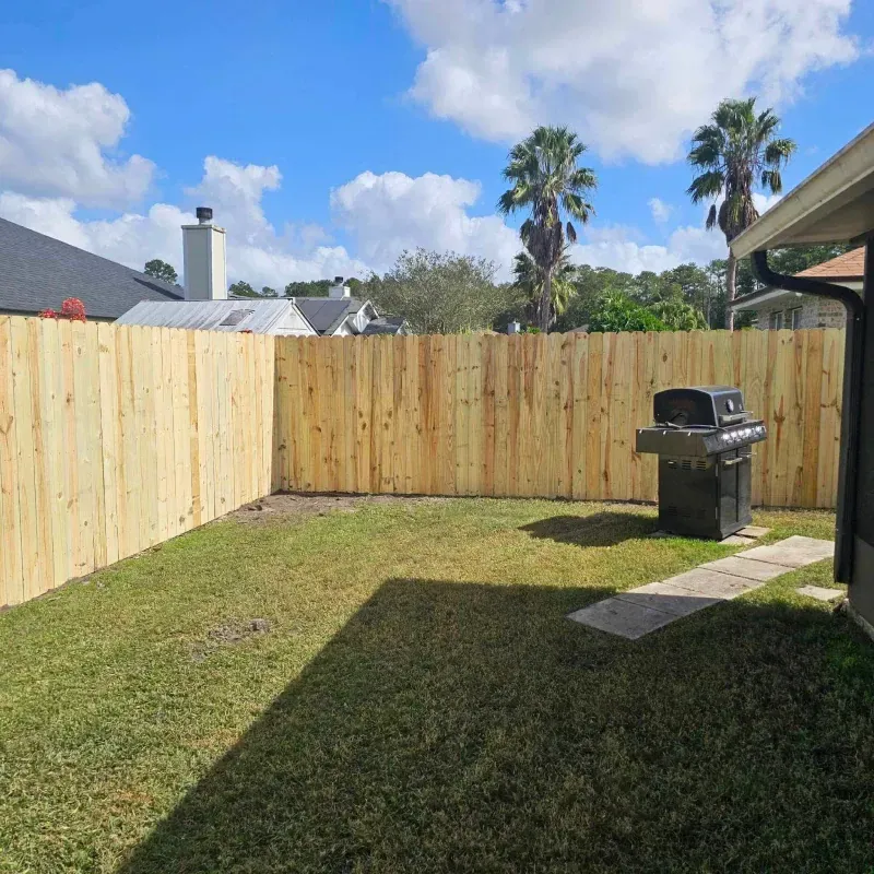 Wooden fence encloses a green lawn with a grill and palm trees under a blue sky with clouds.