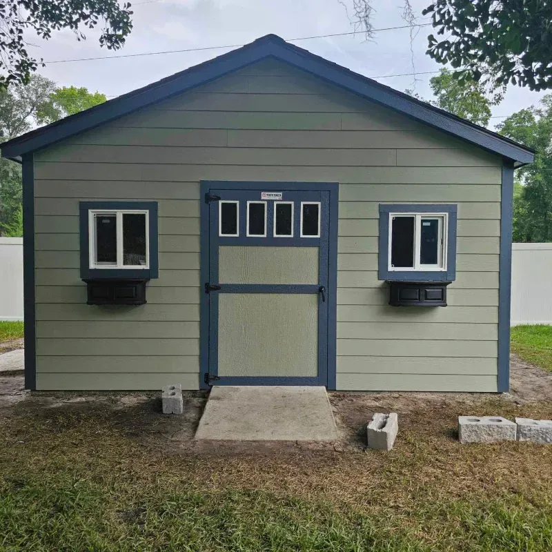 Green and blue shed with two windows, a door, and a concrete pathway on a grassy lawn.