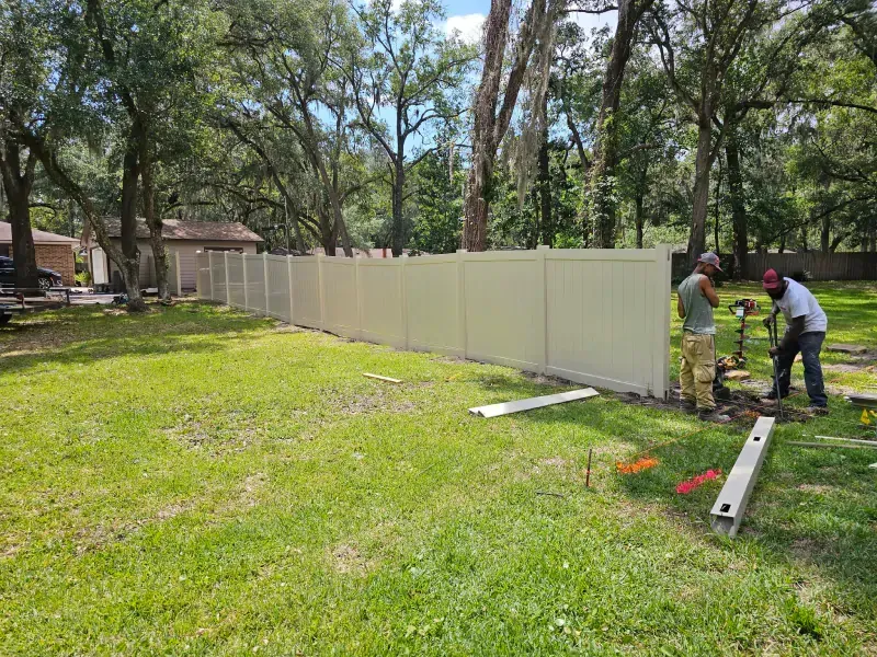 Two workers installing a tan vinyl fence in a grassy yard, with a partly-built fence in the background.