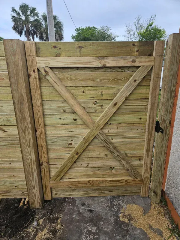 Wooden gate with an X-shaped brace, set in a wood fence with palm trees in the background.