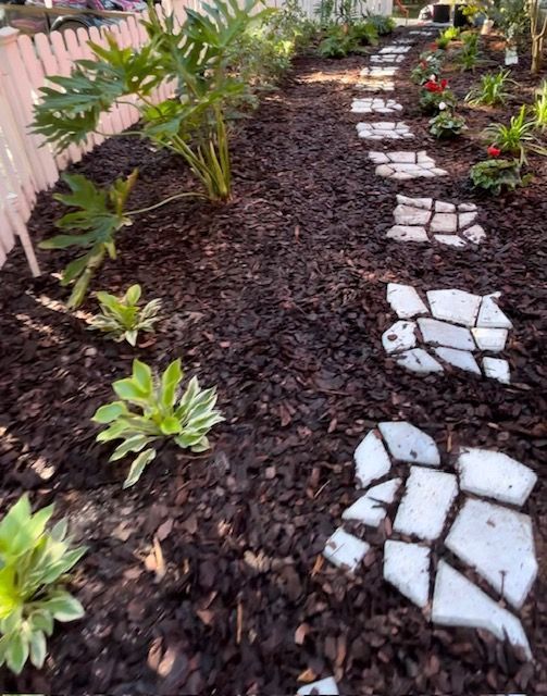 Garden pathway with stepping stones surrounded by mulch and plants. Pink fence on left.