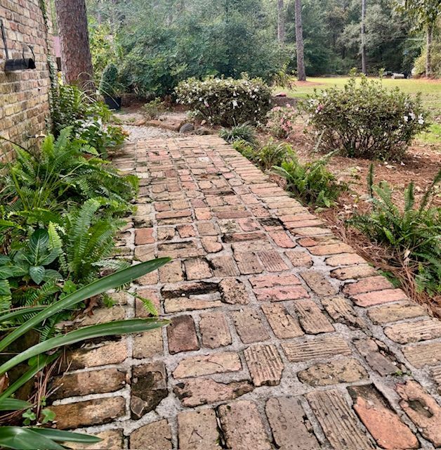 Brick pathway bordered by greenery, leading towards a wooded area.