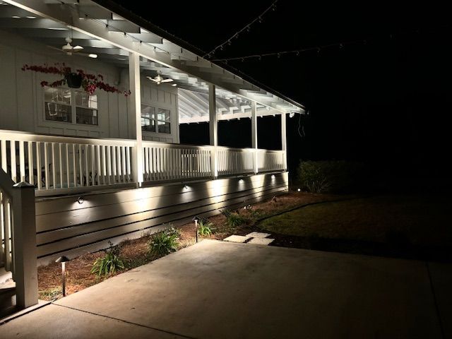 Nighttime view of a house porch with illuminated lights. Dark sky, light-colored porch, and landscaping.