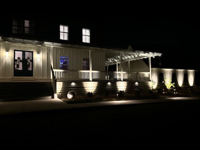 Lit-up white house at night with landscape lighting illuminating the porch, steps, and surrounding greenery.