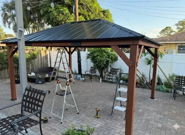 Brown wooden gazebo with black metal roof on brick patio, with ladders and chairs.