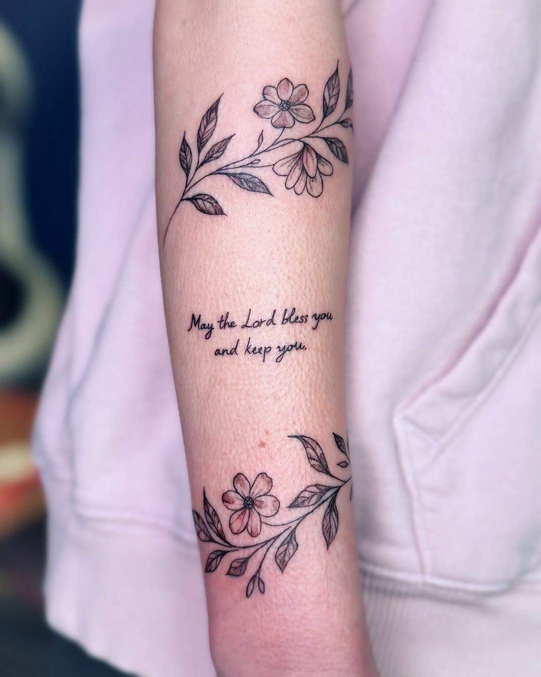 A forearm tattoo featuring two floral branches with the script text 