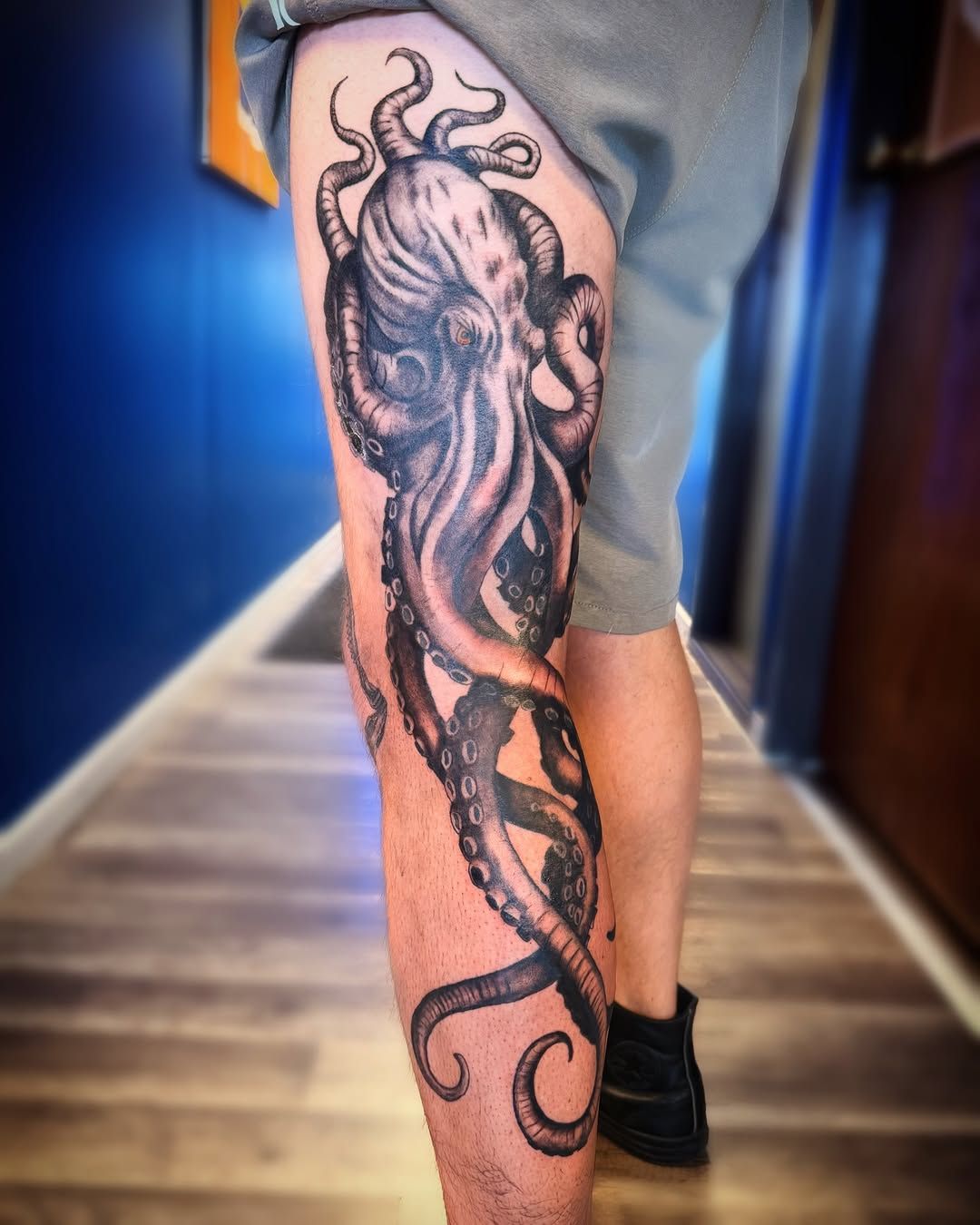 Leg tattoo of a stylized octopus on a person's calf, shown indoors on a hallway floor.