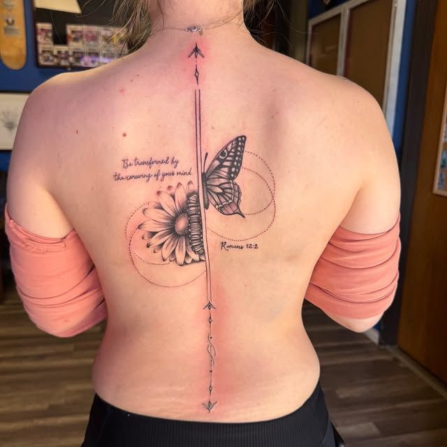 A person’s back with a tattoo featuring a central vertical line, a daisy, a butterfly, and a script quote.