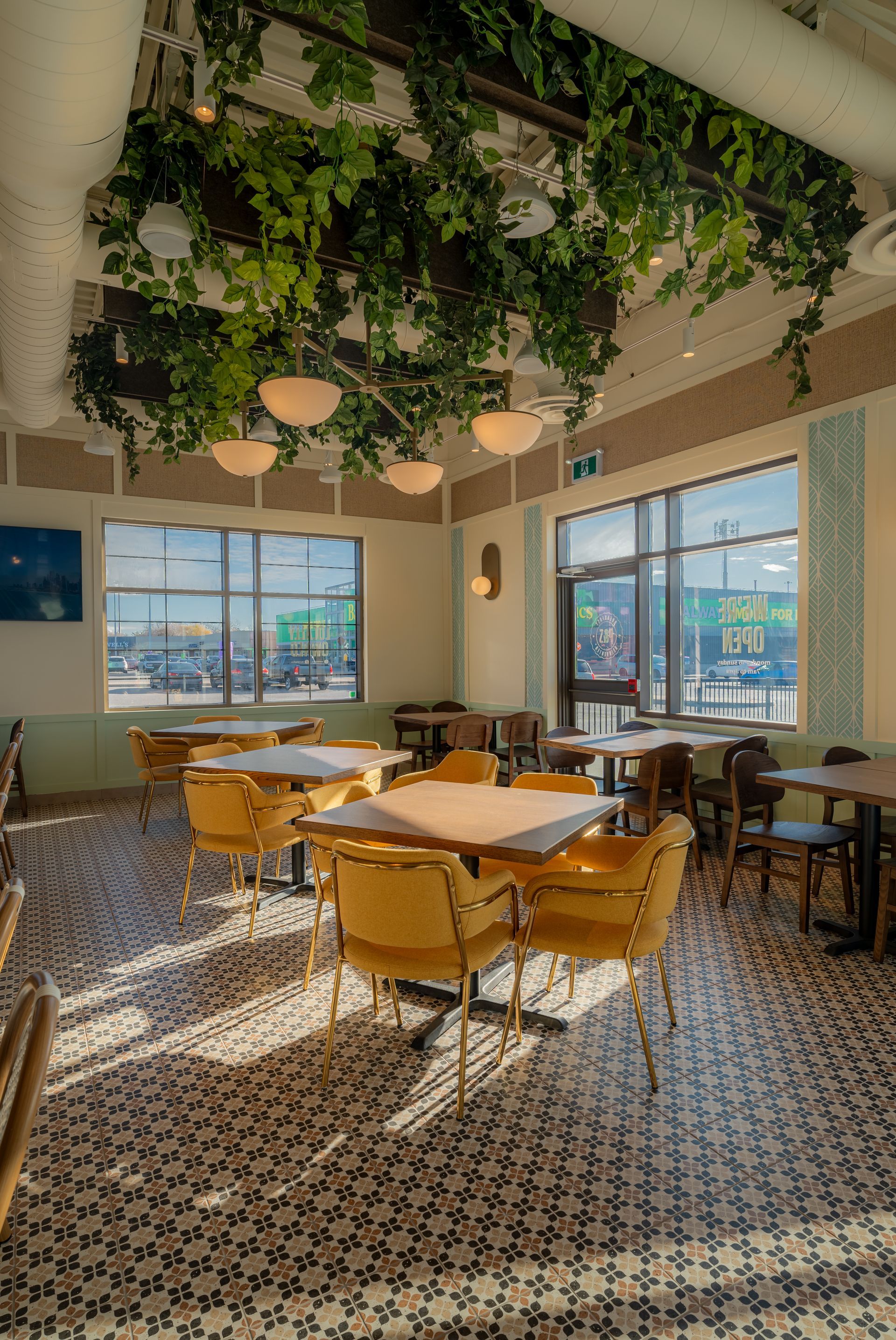 Interior of a restaurant with yellow chairs, square tables, and a patterned tile floor. Vines hang from the ceiling, and sunlight streams through the windows.