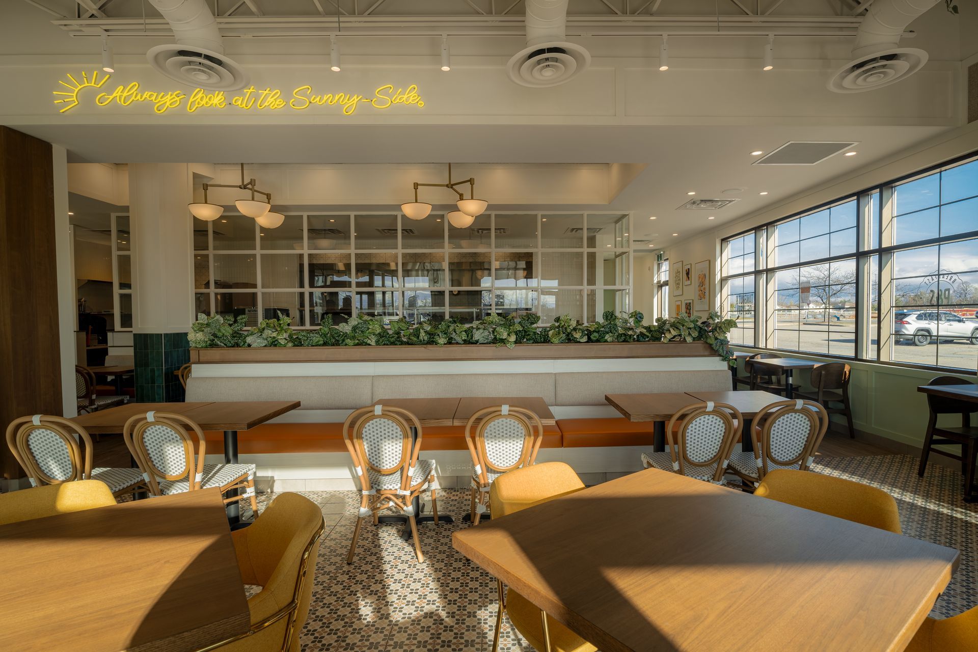 Interior of a bright restaurant with yellow chairs and tables. A long booth with greenery sits against a wall of windows.