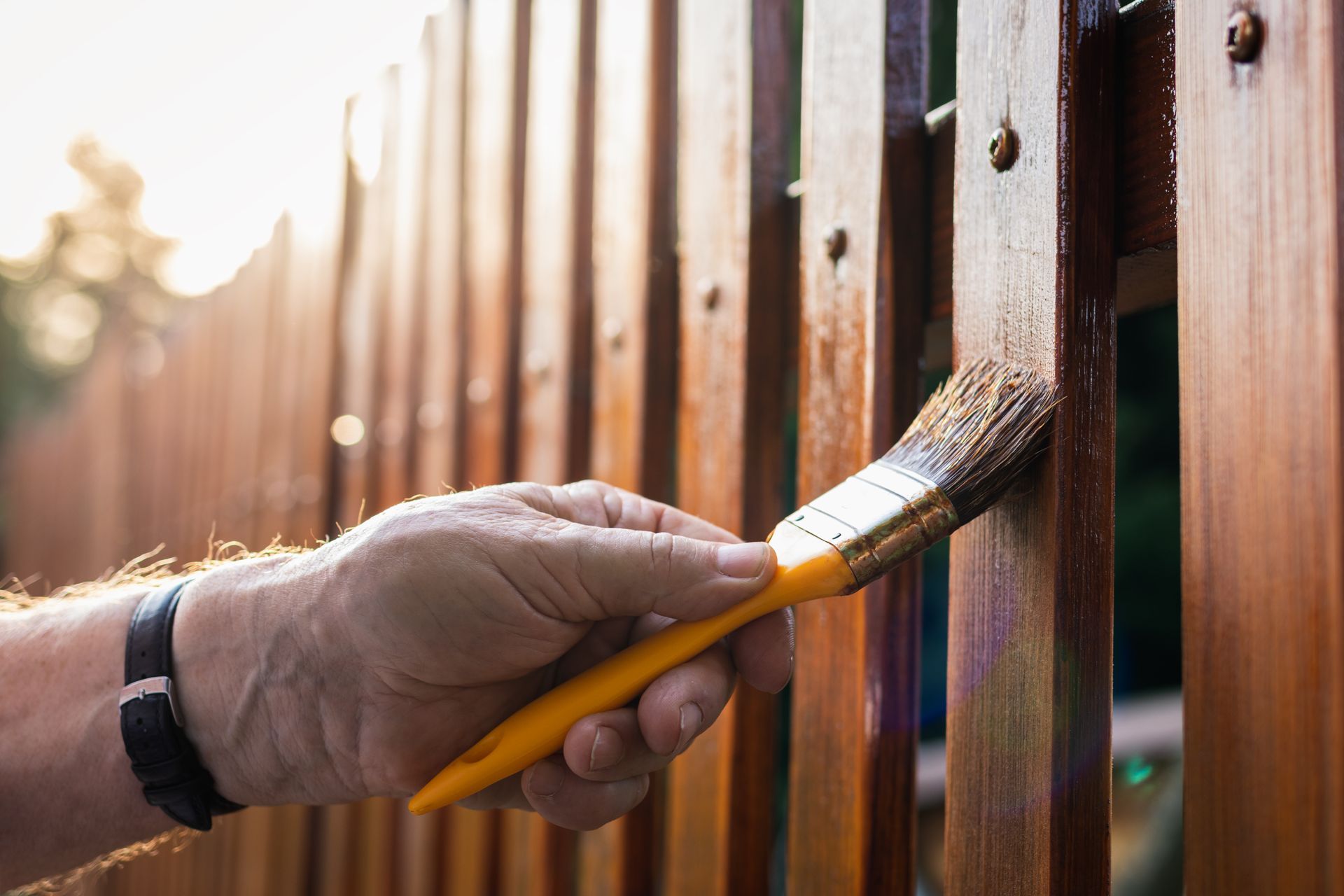 Hand painting a wooden fence with a yellow brush in warm sunlight