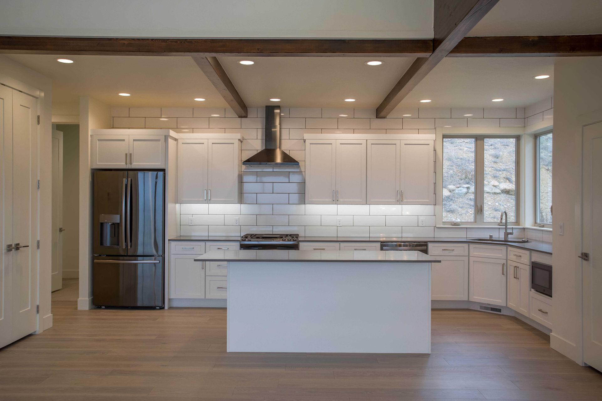 Modern white kitchen featuring an island, stainless steel refrigerator, white cabinets, and wooden ceiling beams.