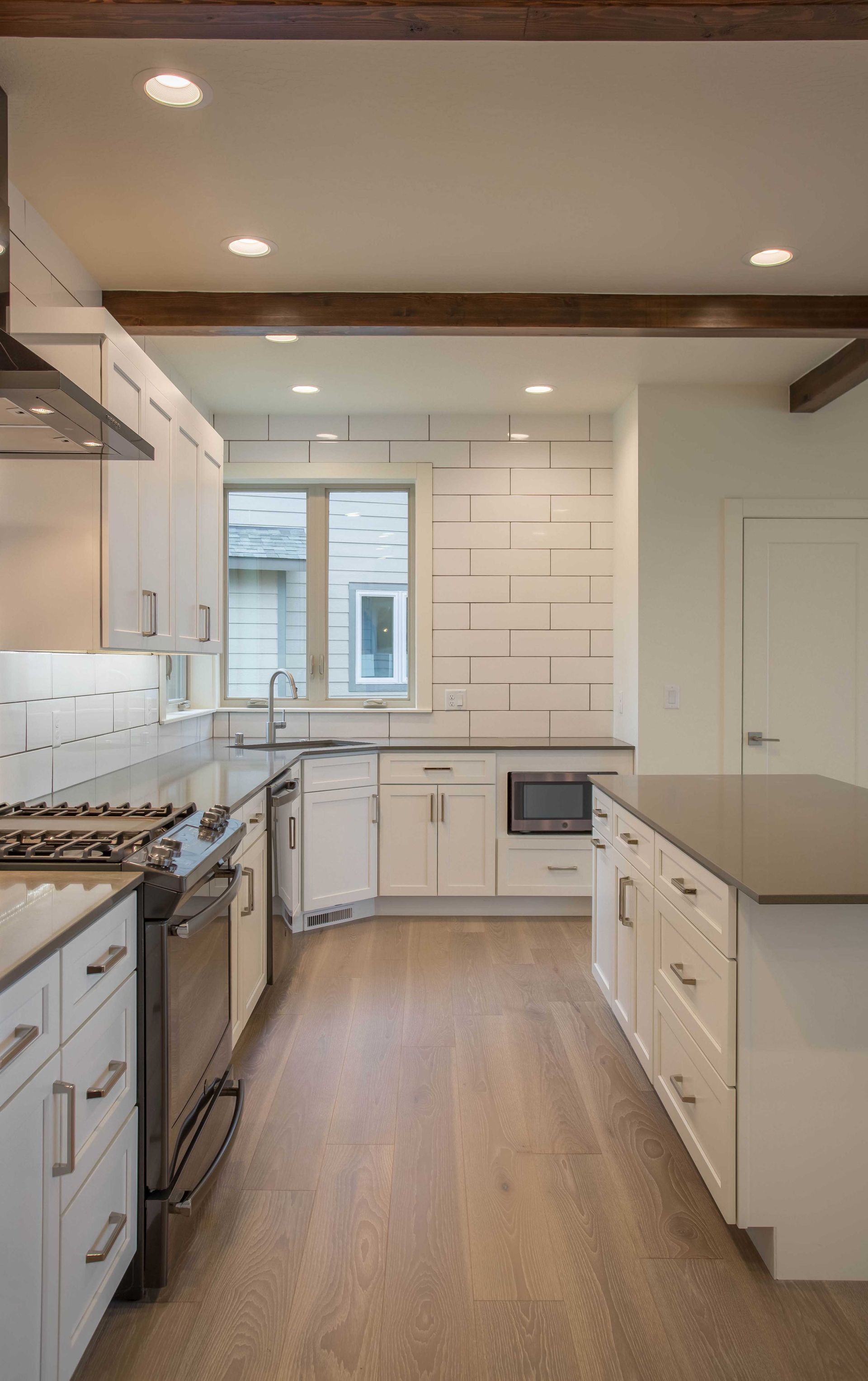 Modern white kitchen featuring a large island, wood flooring, and a tiled backsplash under wooden ceiling beams.