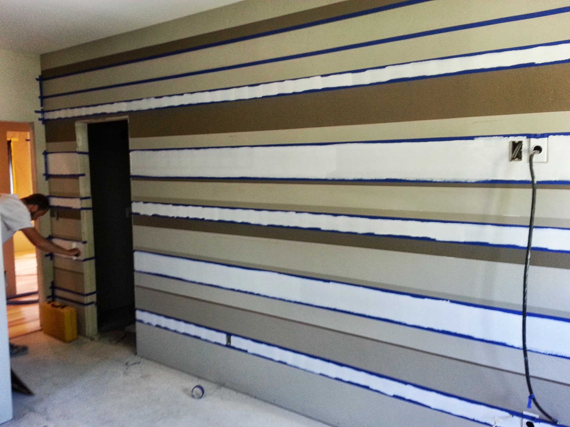 A person painting horizontal beige and white stripes on a room wall using blue painter's tape to mark the edges.