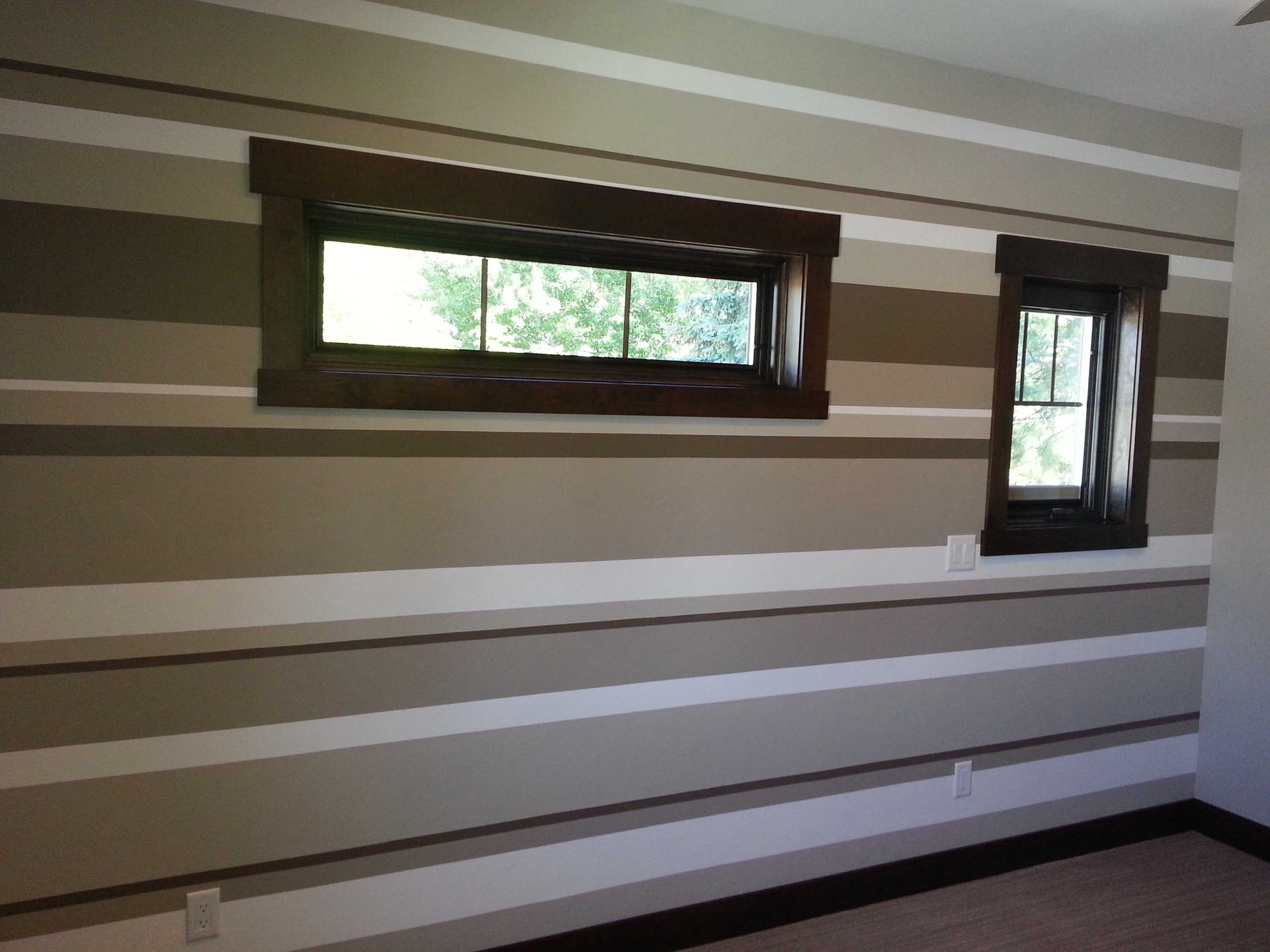A room wall painted with horizontal stripes in white, beige, and brown, featuring two rectangular windows with dark frames.