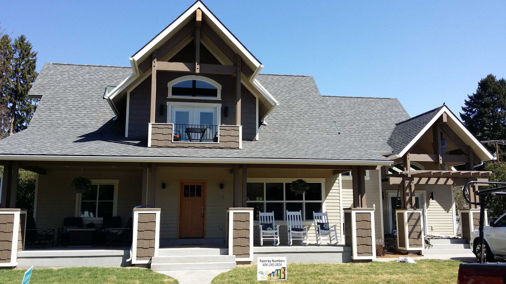 A two-story tan home with a large front porch, shingled roof, balcony, and brown trim on a sunny day.