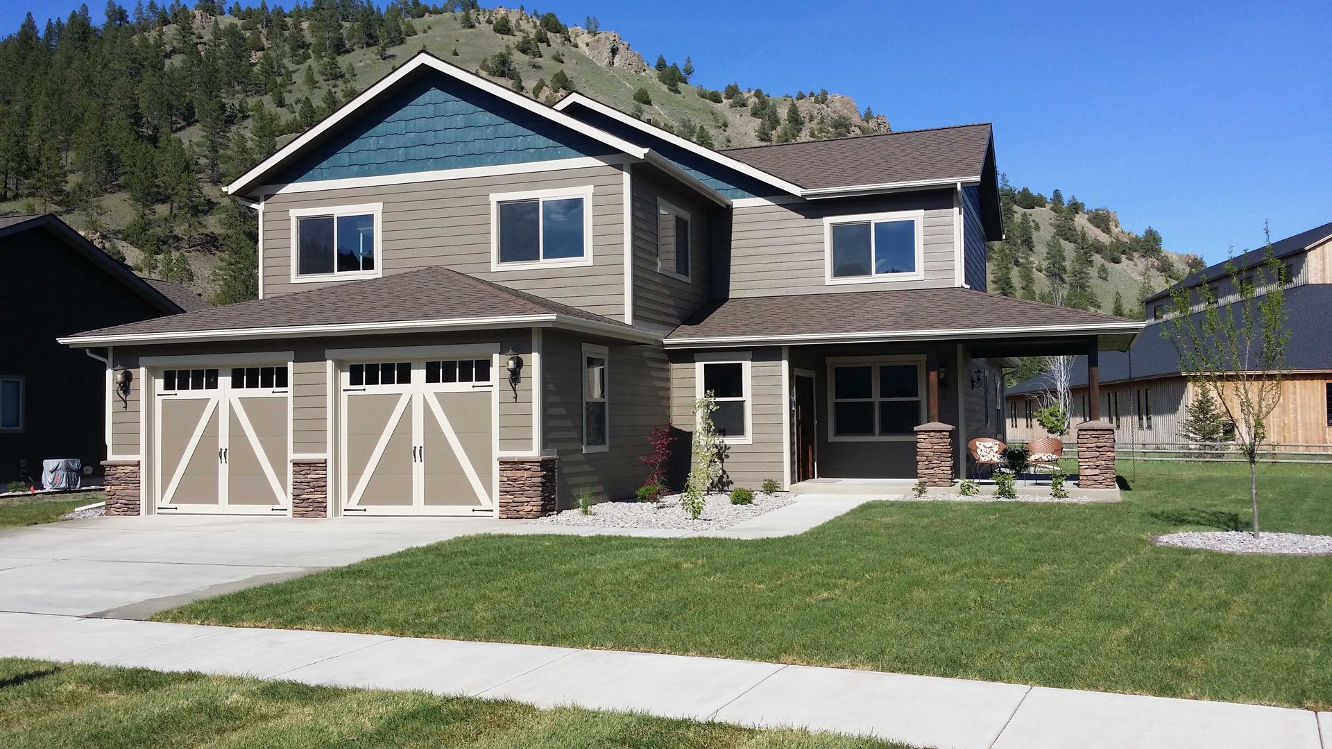 Two-story taupe house with a two-car garage, covered porch, and blue gable accent against a mountainous backdrop.