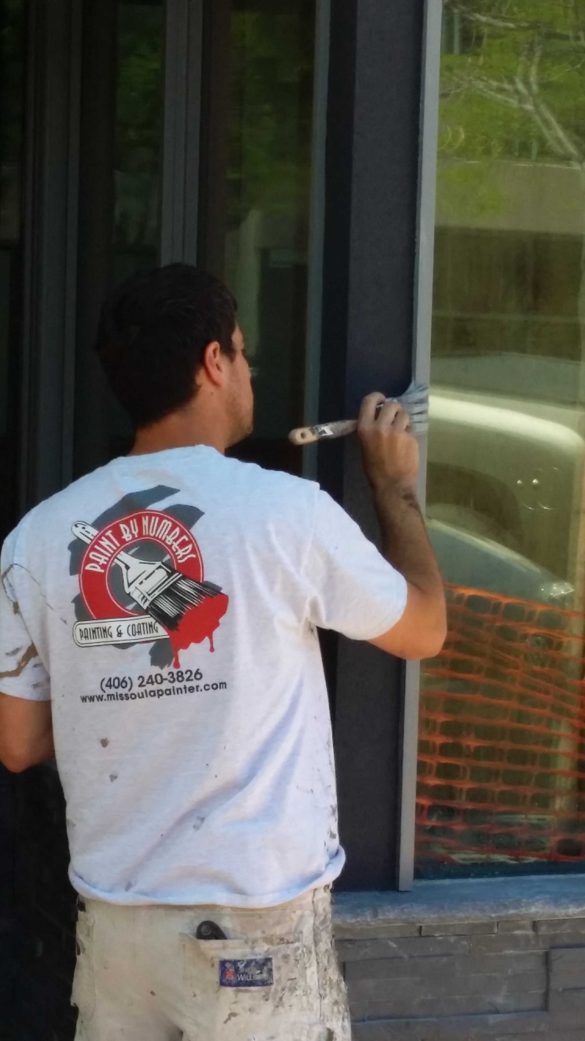 A professional painter wearing a branded t-shirt uses a brush to paint a dark window frame.