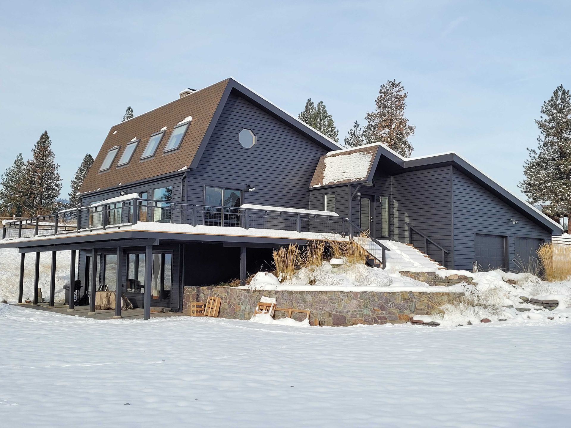 A dark-sided two-story house with a large deck and skylights, surrounded by snow-covered ground and pine trees.