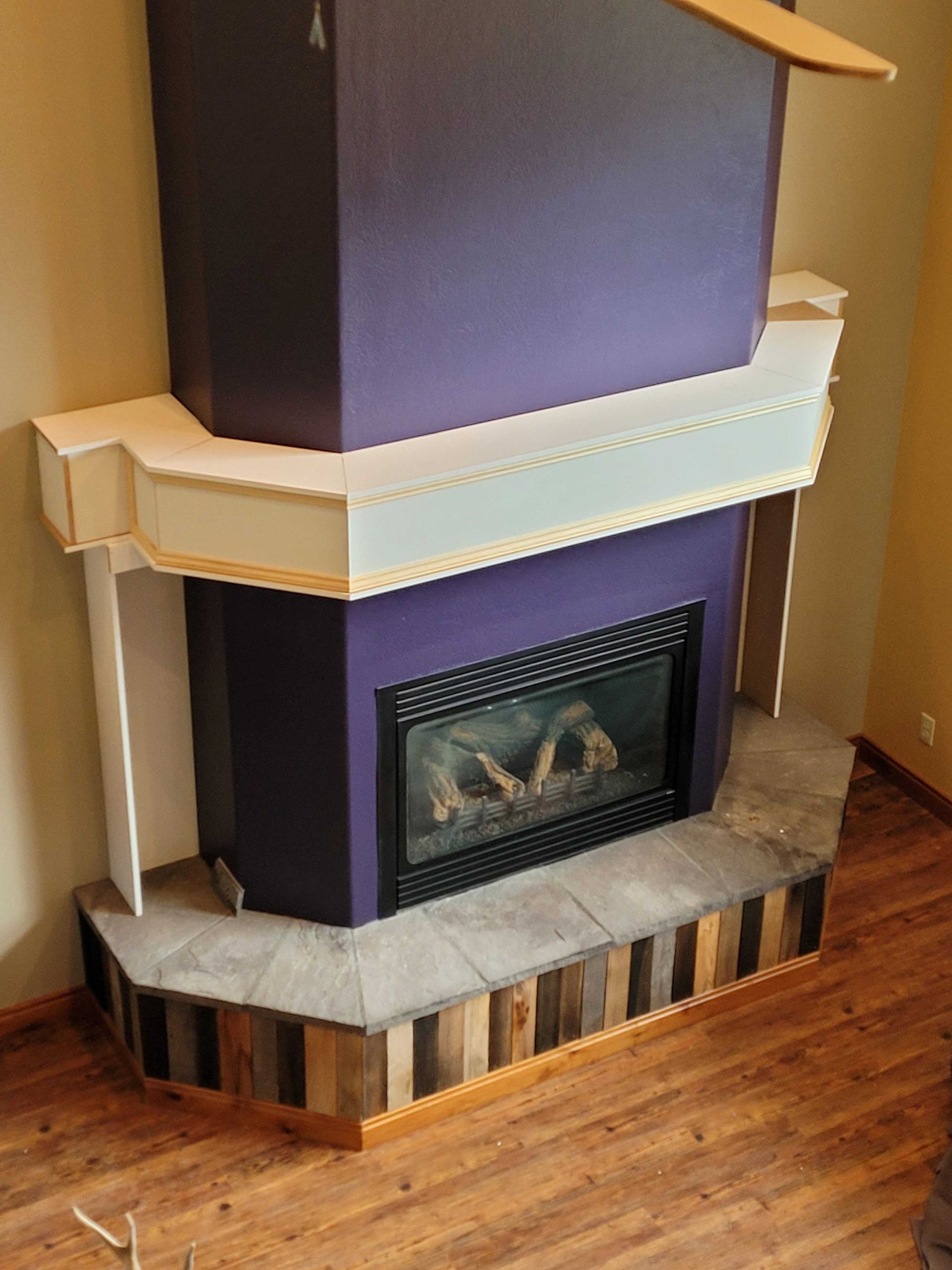 A corner fireplace featuring a purple chimney, a white mantle, stone tiling, and a wooden base with vertical planks.